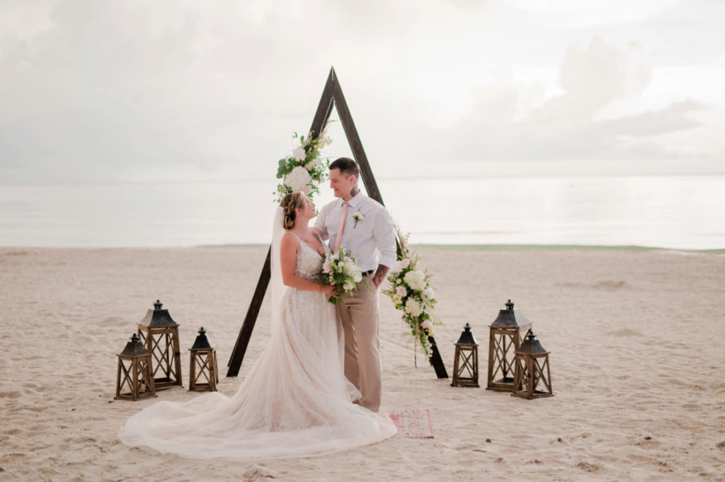 Boho Beach Wedding Package, Bride and groom standing on the beach in front of dark A-frame arbor with fresh flowers and wooden lanterns