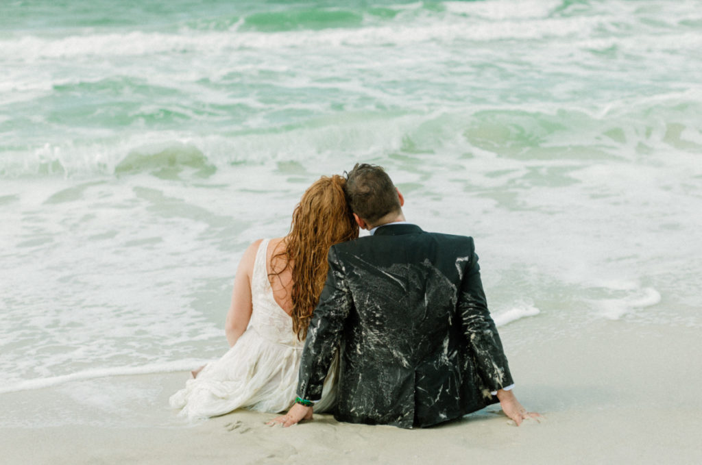 Bride and groom sitting on the sand as the waves cascade along the beach.